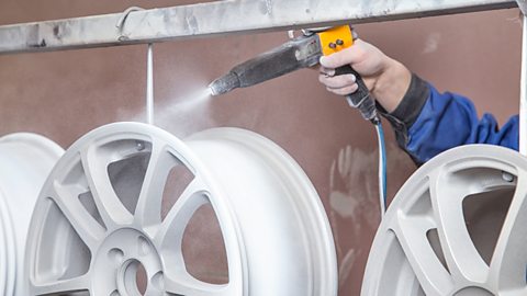 A metal car wheel rim is suspended from a bar while a hand powder coats it white.