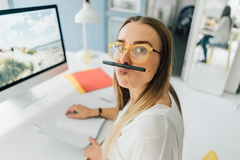 A girl sits at a desk and jokes around making a moustache with a pencil
