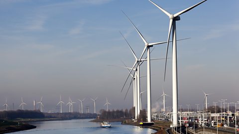 A collection of wind turbines are shown in place on a riverside generating electricity.