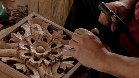 Hands use a chisel and hammer to carve a flowered pattern into a piece of timber.