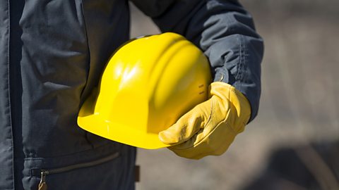 A gloved hand holds a bright yellow hard hat against dark blue overalls.