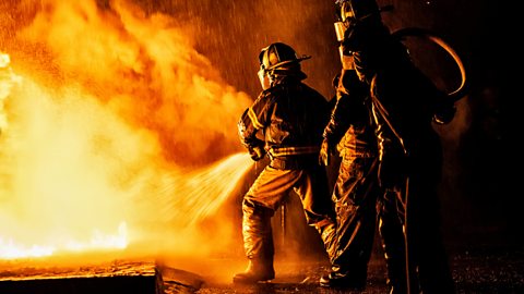 Two firefighters fight off a large blaze with water from a hose.