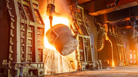 Molten steel being poured from a large metal cauldron, causing sparks, in a steel production plant.