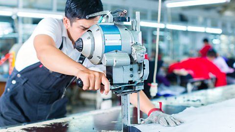 A worker wears a chainmail glove as he operates a large band knife to cut a fabric sheet.