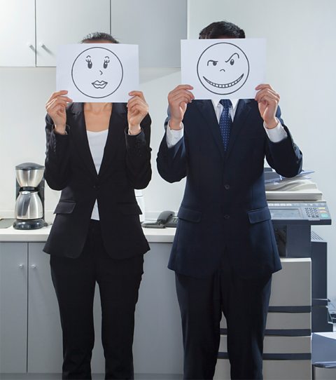 A businesswoman and businessman stood side by side holding smiley faces on paper.