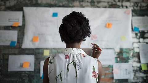 A teacher looking at a board with post it notes on it. The teacher has a pen in their hand.