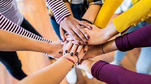 Five pairs of hands on top of each other, the hands belong to a group of people who are standing in a circle.