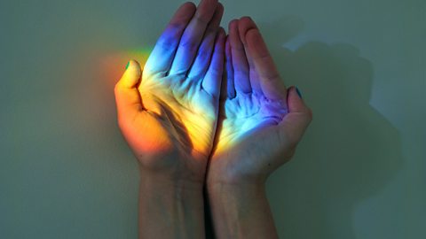 A pair of hands close together on a surface with their palms facing up. There is a rainbow light shining onto the palms.