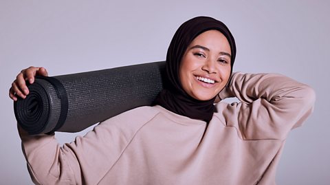 A person standing with a yoga mat on their shoulders. They are smiling and are wearing a pink sweater and a black headscarf. 