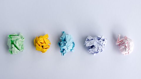 Five scrunched up pieces of paper on a white desk. The paper is colourful, green, yellow, blue, purple and pink.