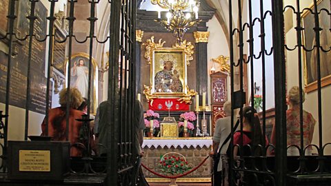 People worshipping at a small altar with a depiction of The Virgin Mary, seen through a partition