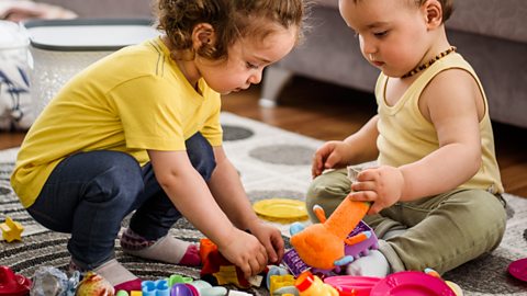 A little girl playing with toys while her baby brother watches.