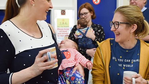 A mum holding a baby, chatting to another mum over a cup of tea.