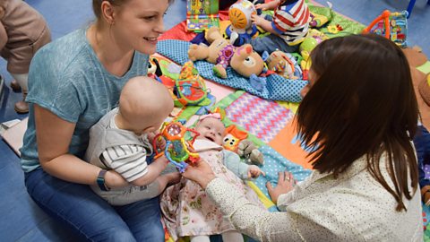 Two mums chatting to each other at a play group, with a baby boy and girl on a mat with toys.