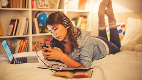 A student laying ona bed listening to music on their phone. They are wearing headphones and has a work book and laptop open on the bed.