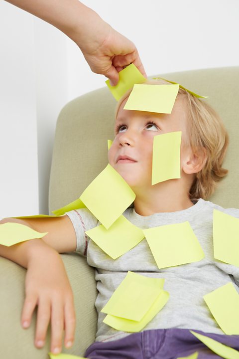 A child sitting in a green chair with post it notes stuck all over their face and body.