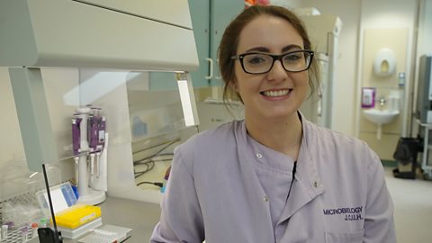 Hannah sitting in a lab wearing a lab coat and smiling to camera.