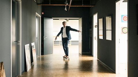 A man in smart clothes skateboarding down an empty school corridor. 