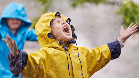 Two children laughing and playing in the rain. One is wearing a yellow waterproof jacket and the other is wearing a blue jacket.