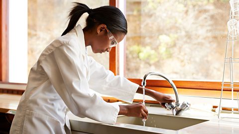 A young person dressed in a labcoat and goggles filling a glass tube with water from a tap.