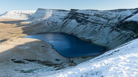 Golygfa o lyn wrth ymyl mynydd serth, wedi'i orchuddio gan eira