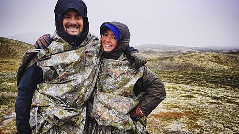 A man and a woman wearing camouflage onesies on a similar looking mountainous background. 