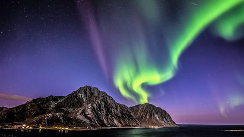 Green lights in the night sky over a huge rock and small town. Wide shot.