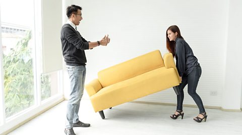 A woman struggles to move a couch and glares at her partner while he stands and cheers her on.