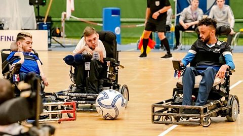 Three men in powerchair wheelchairs with a football in the middle.