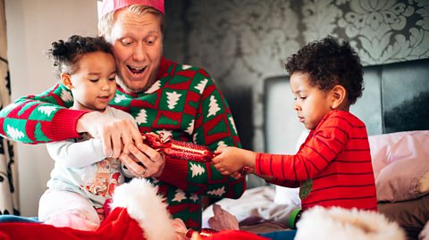 Two young children play with a red Christmas cracker, pulling one side each, while their dad helps them. 