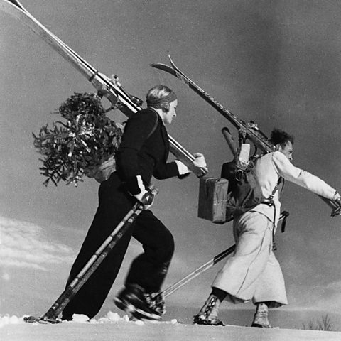 German couple climbing up a snow hill with skis, a christmas tree and parcels (1937)