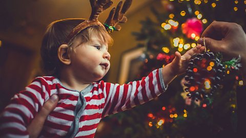 Toddler wearing reindeer headband being supported by adult to hold a Christmas decoration