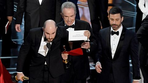 Three men standing in front of a microphone, one is holding a piece of paper and another is holding a trophy.