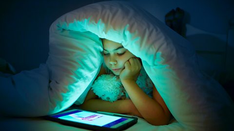 A kid hidden under the covers looking down at a tablet, the screen's glow illuminating her face