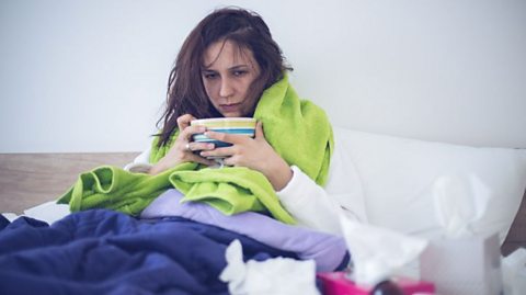 An ill woman in bed with a blanket, surrounded by used tissues, is eating soup.