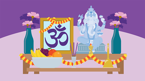 A table with items on it including vases of flowers, a bowl of fruit, incense sticks in a silver pit, a golden bell, a statue of the Hindu god Ganesha, the elephant-headed god, and a framed picture.