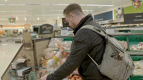 A little girl holding up a carrot to her dad as she rides in a supermarket trolley.