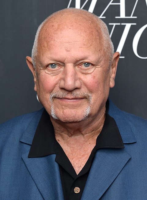 Theatre practitioner Steven Berkoff wears a textured blue suit over a black shirt at a red carpet event.