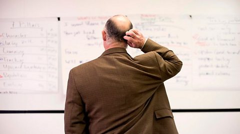 A teacher is stanging in front of a whiteboard and scratching the back of his head.