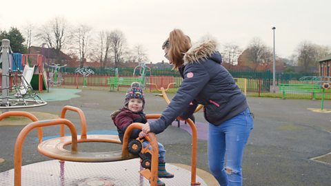 Child sitting on merry go round while mum holds on.