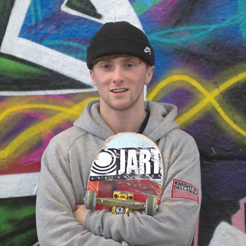 Alex DeCunha is standing in front of a wall of graffiti holding his skateboard.