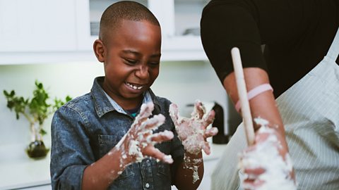 A child looks at his hands which are covered in dough and he's smiling 