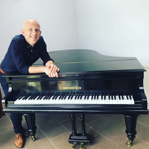 Danny Lane standing by his piano at home and smiling.