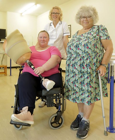 Miriam stands next to a physio and Angela (sitting in front of the two) in Miriam's Big Fat Adventure, Angela is holding her new prosthetic leg