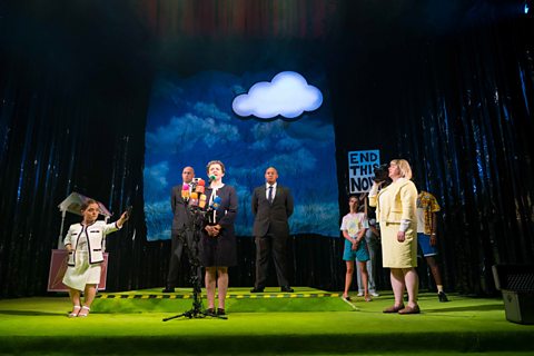 The cast of the play Pity stand on a colourful set featuring a fake grass floor and a large cartoon cloud