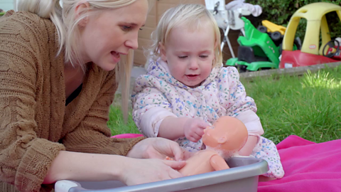 A mum and her little girl talking while the little girl points at the nose of a plastic doll.