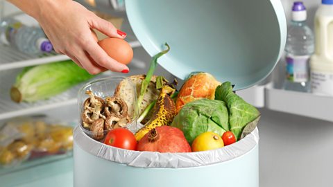 A lady puts fruit and veg in a bin with an open fridge door behind her 