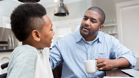 Dad talking to son sitting at a kitchen table. The dad is holding a cup of tea and wearing a blue shirt.