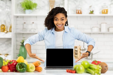 A young woman stands behind her laptop with food either side of the laptop, suggesting she bought the food online