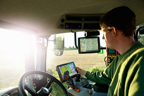 A farmer sits in a tractor while looking at small screens (highlighting he's using technology to farm with)